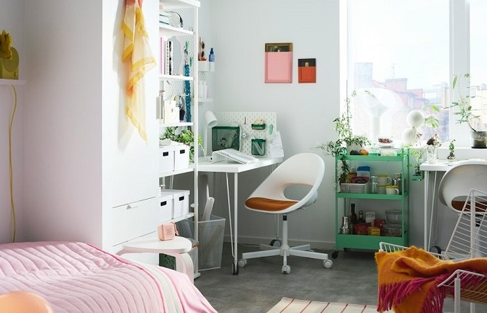 A brightly lit college student's room featuring a bed with pink striped bedding, a white shelving unit with storage boxes, a small white desk with a white swivel chair, and a green trolley holding plants and tableware. 