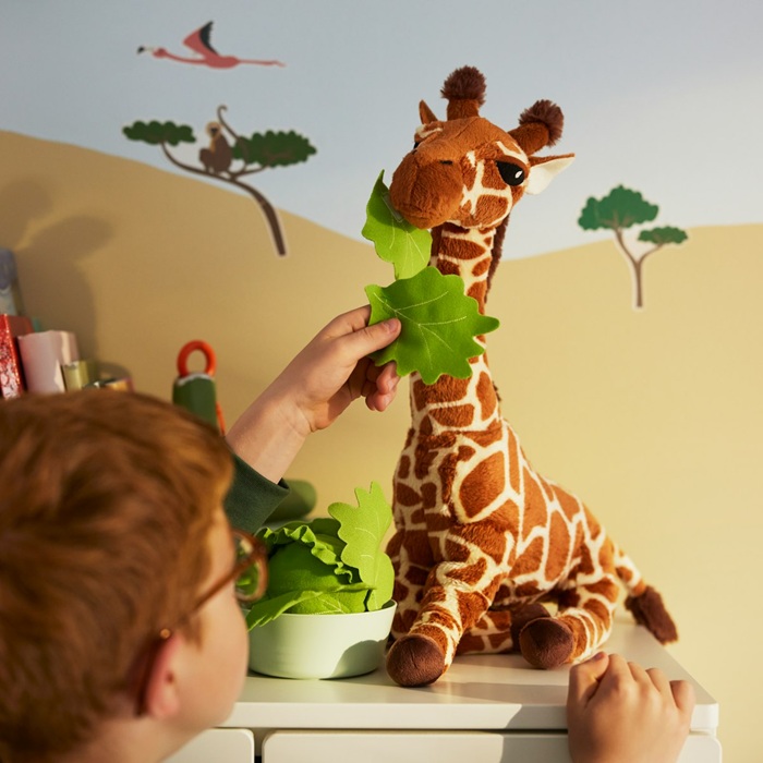 A child feeds toy leaves to a SANDLÖPARE soft toy giraffe in a play area with a savannah-themed wall.