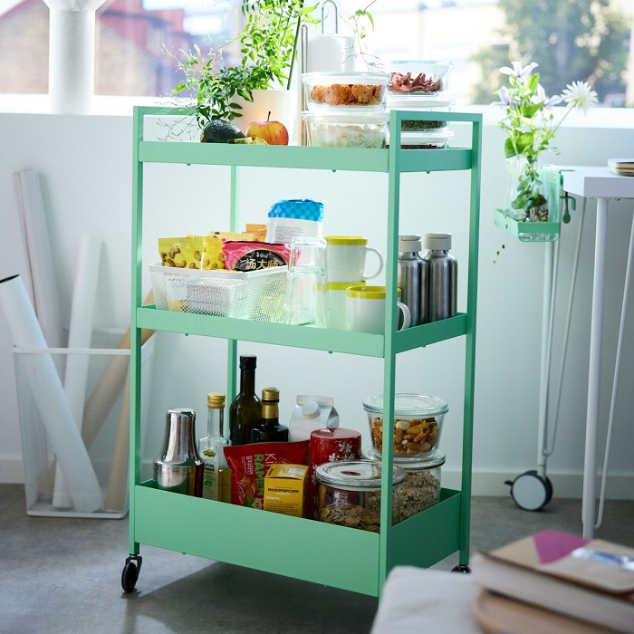 A light green NISSAFORS trolley in a bright room, stocked with various snacks, jars, and kitchen items across its three shelves. 