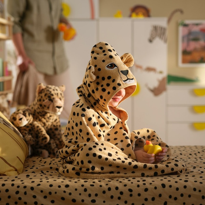 A child in a SANDLÖPARE towel with hood with cheetah pattern sits on a bed surrounded by soft toy cheetahs and a rubber duck.