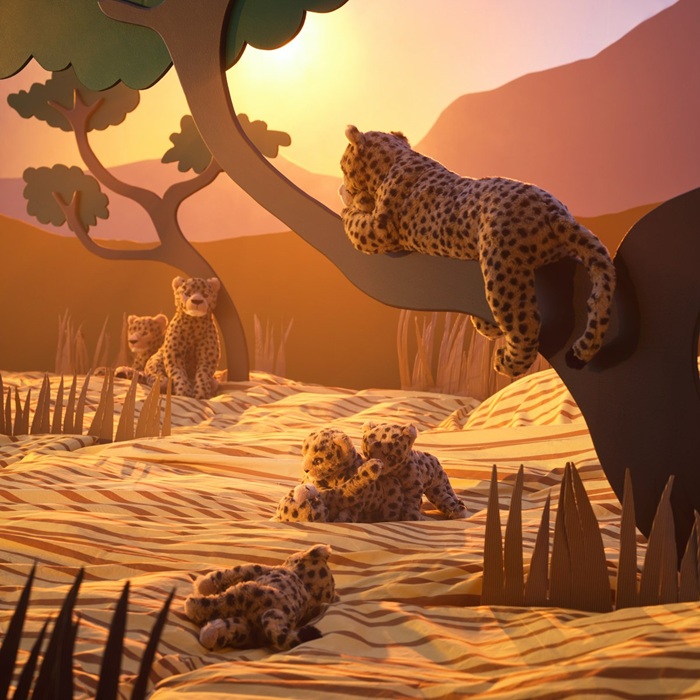 SANDLÖPARE soft toy cheetahs rest on the ground and in a tree in a sunlit savannah landscape made of cardboard.