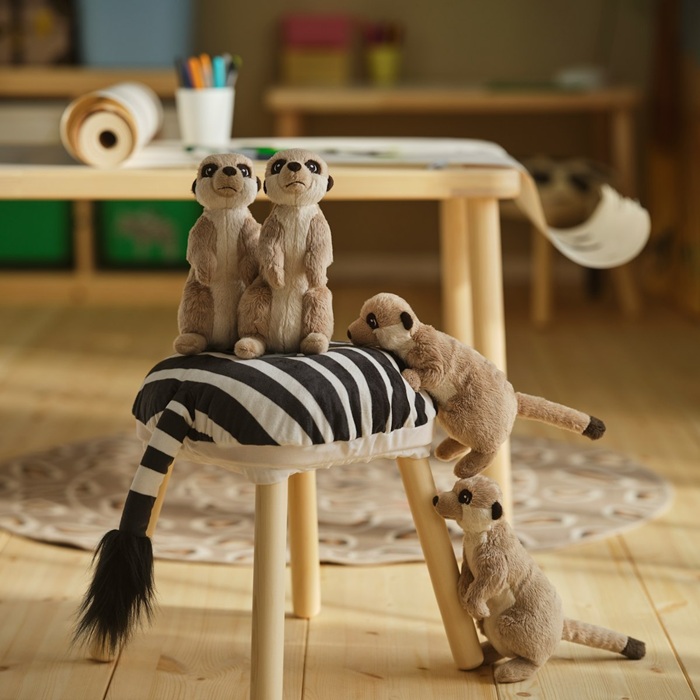 Divas SANDLÖPARE mīkstās rotaļlietas surikati bērnistabā, sēdus noliktas uz taburetes ar zebras raksta pārklāju. Vēl divas it kā rāpjas augšup pa taburetes kāju.