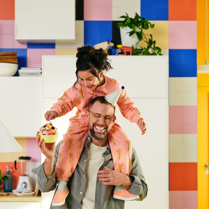 Parent carrying a smiling child on their shoulders in a colourful kitchen.
