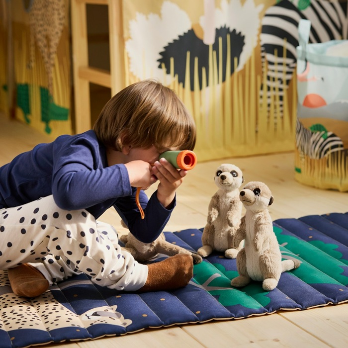 A child looks through a toy telescope at SANDLÖPARE meerkat soft toys on a play mat in a savannah-themed room.