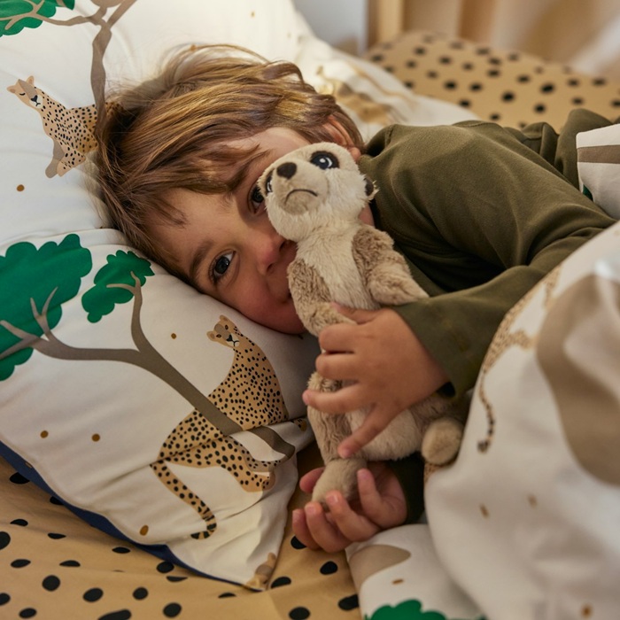 A child cuddles a SANDLÖPARE meerkat soft toy while lying in bed with savannah-themed bedding.