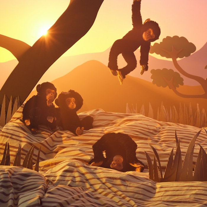 A black SANDLÖPARE soft toy chimpanzee hangs from a tree, while others sit on the ground in a sunny savannah landscape.
