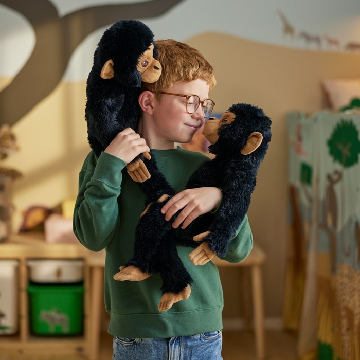 A child smiles while holding two black SANDLÖPARE soft toy chimpanzees, one on each shoulder, in a savannah-themed children’s room.