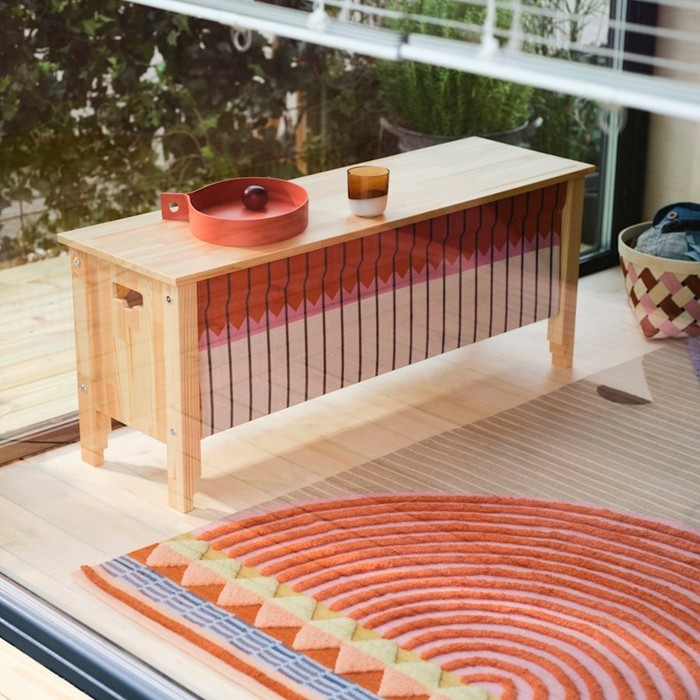 A wooden OMMJÄNGE bench with storage features a textile front showcasing weaves in red, pink and white tones. The bench is placed against a glass wall.