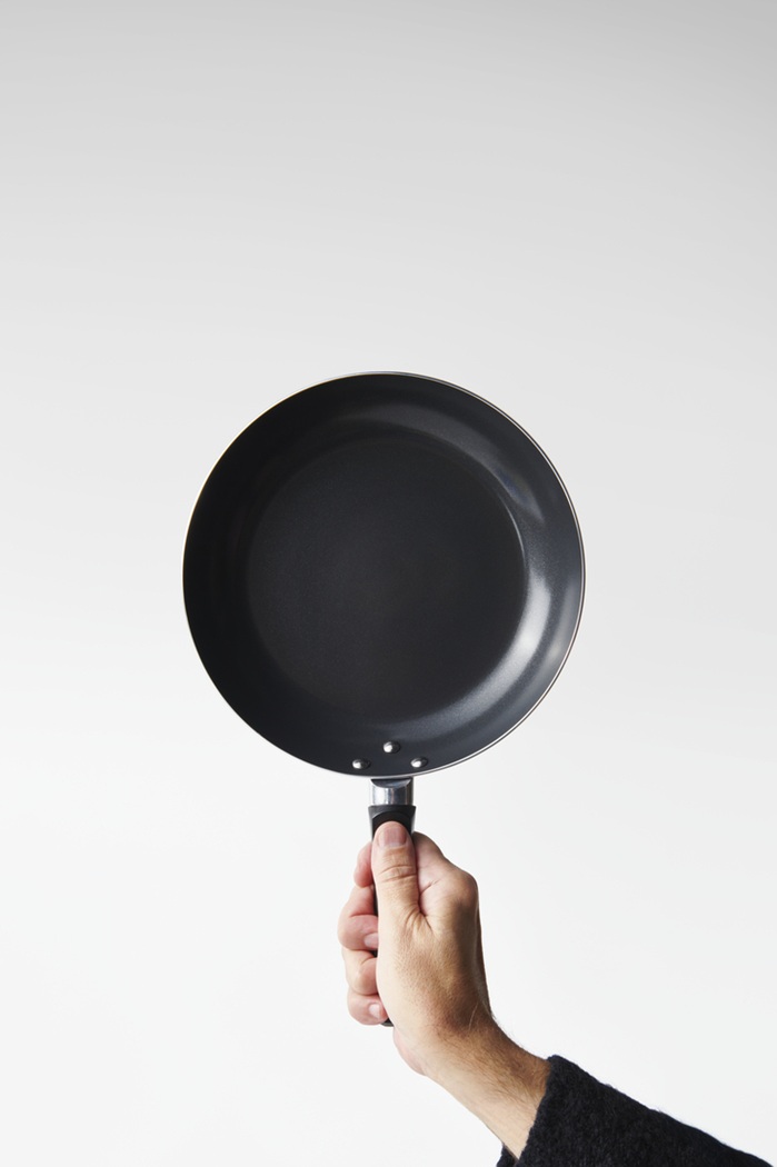 Hand holding up the TAGGHAJ black frying pan against a plain white background.