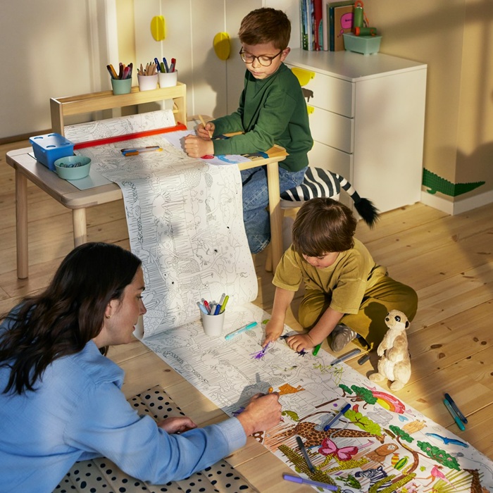 Two children and a parent colour together on a large roll of savannah-themed colouring paper spread on the floor.