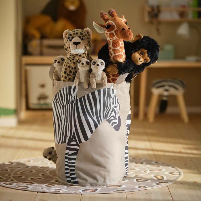 A SANDLÖPARE storage bag filled with soft toy animals, including a giraffe, cheetahs and a chimpanzee, sits on a patterned rug.