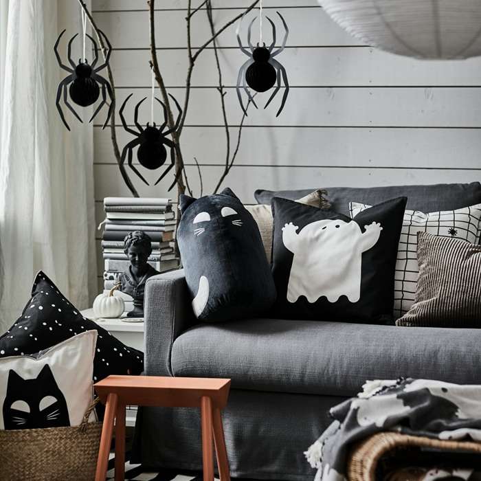 A cosy seating area with a grey sofa adorned with KUSTFYR cushions and throws in spooky Halloween-themed designs, including a ghost and a cat face. In the background, black spiders are hanging from tree branches.