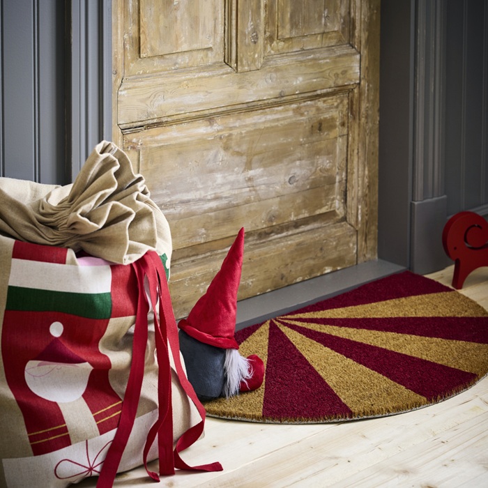Праздничный уголок у деревянной двери с красно-золотым круглым ковриком, маленькой фигуркой рождественского гнома и упакованными подарками.