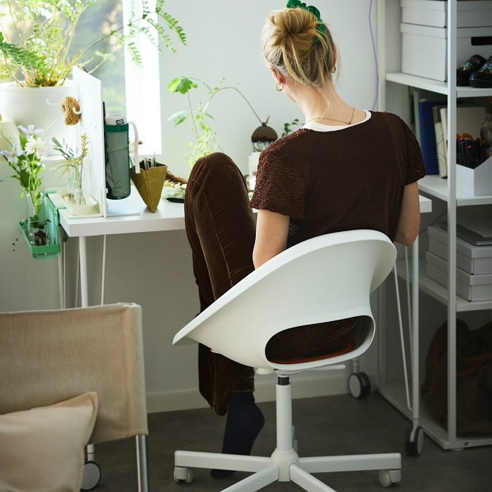 A person sitting on a white LOBERGET swivel chair at a desk by the window, with their legs folded up on the seat. 