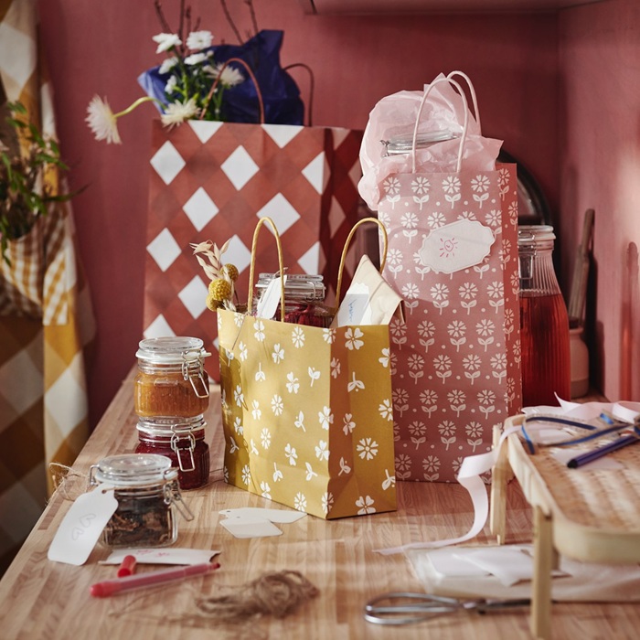 Three patterned KÖSSEBÄR gift bags in red, pink and yellow on a wooden table, filled with jars, bottles and small presents. Ribbons, scissors and wrapping supplies are scattered around.