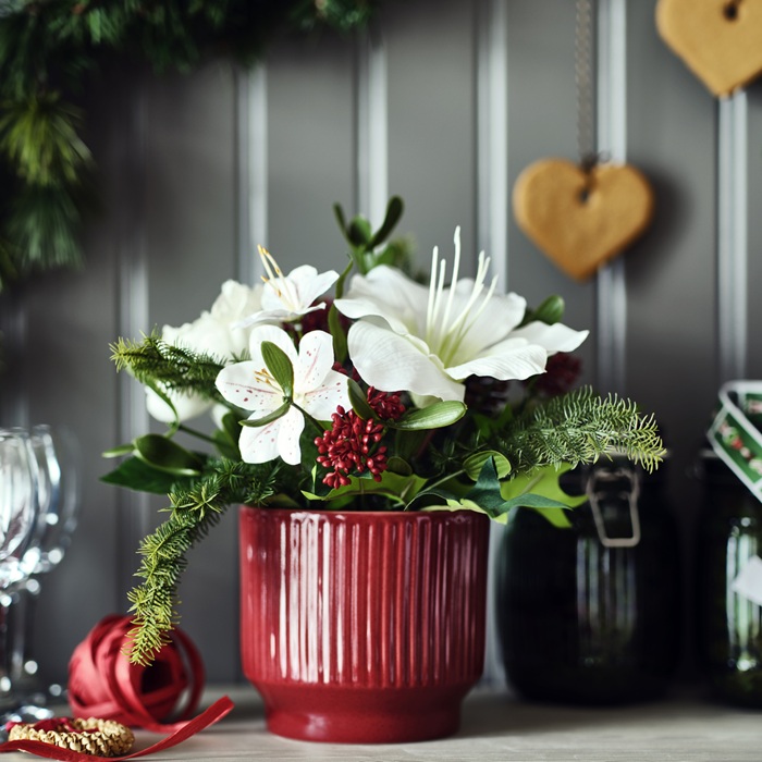 Праздничная композиция из искусственных белых цветов в красном ребристом горшке на фоне темного дерева с подвесными имбирными пряниками в форме сердца.