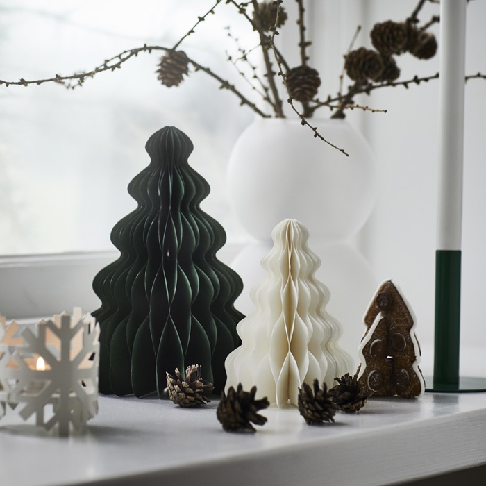 Минималистичная праздничная композиция со складными бумажными елочками темно-зеленого и белого цветов, расположенными на подоконнике рядом с шишками и зимними ветками.