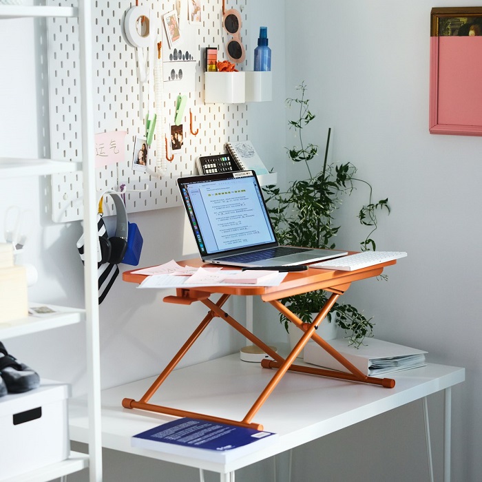 An orange DUBBLA laptop stand is placed on a white desk and holds a laptop. A white SKÅDIS pegboard with hanging supplies is mounted on the wall behind the desk.