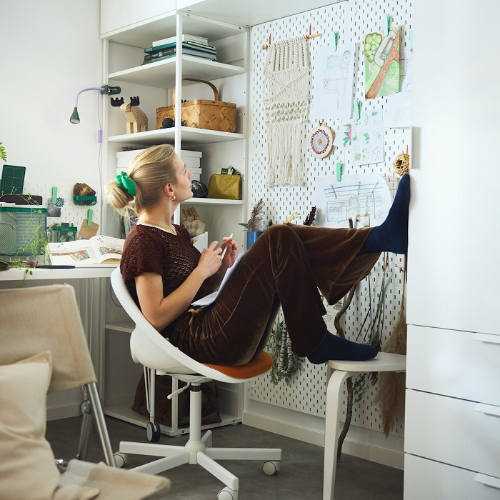 A person sitting on a white swivel chair, looking thoughtfully at a wall-mounted SKÅDIS pegboard. The pegboard is filled with sketches and notes.