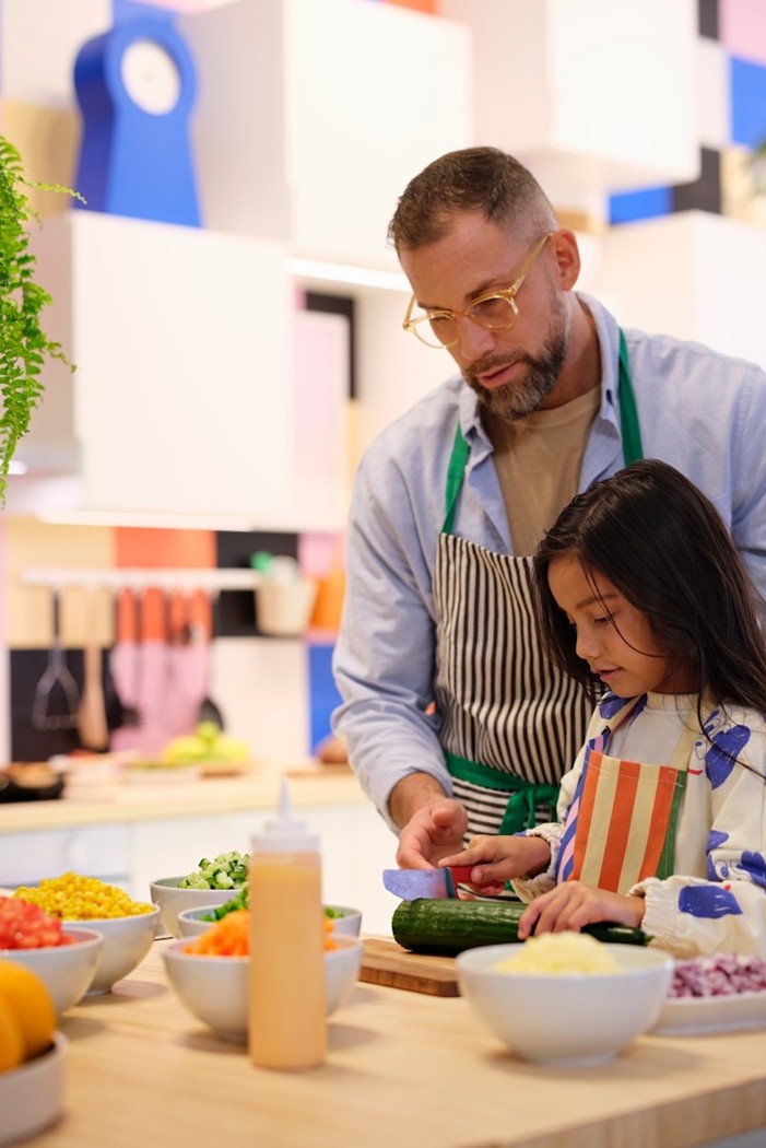 A parent and child stand at the kitchen counter, slicing vegetables side by side, sharing a joyful cooking moment in a bright, playful space.