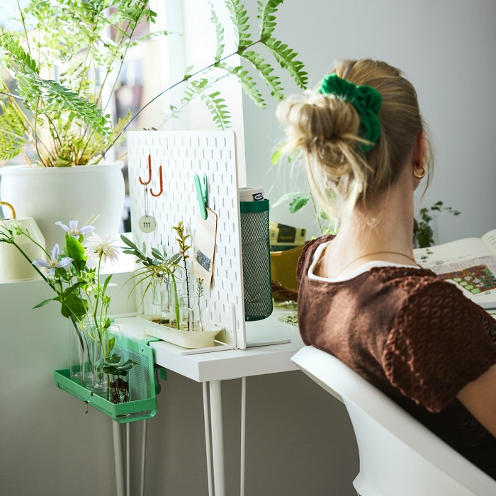A person sitting at a desk surrounded by lush green plants, facing a small SKÅDIS pegboard mounted on the desk.