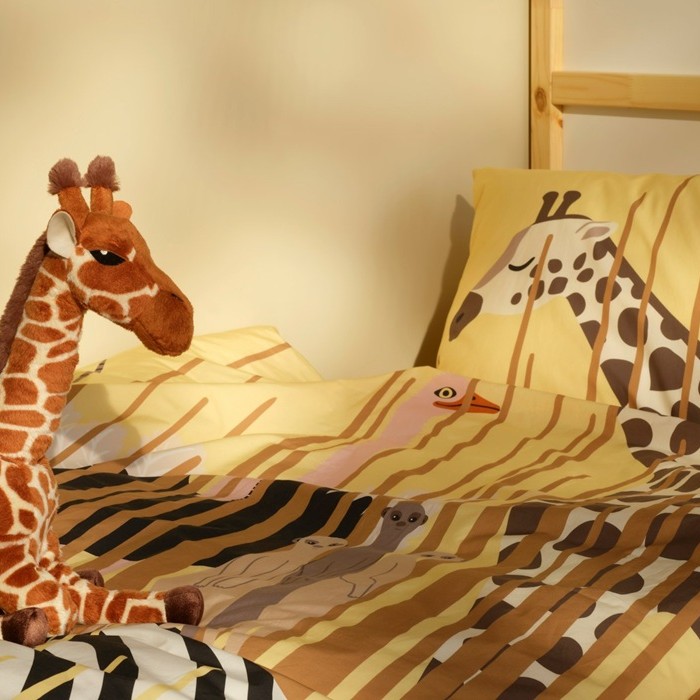 A SANDLÖPARE soft toy giraffe sits on a bed with savannah-themed bedding featuring giraffe patterns.