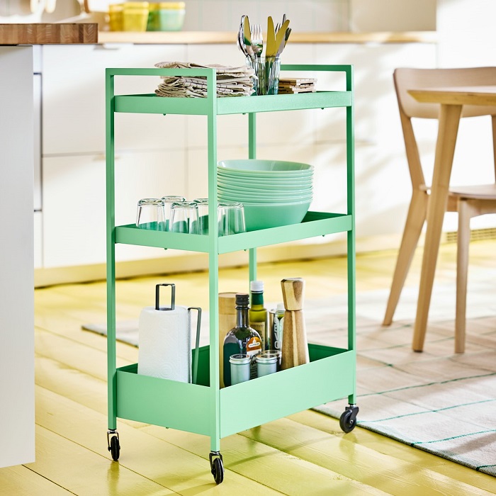 A green NISSAFORS trolley with three shelves, holding glasses, dinnerware and table accessories, is placed in a bright, airy kitchen.