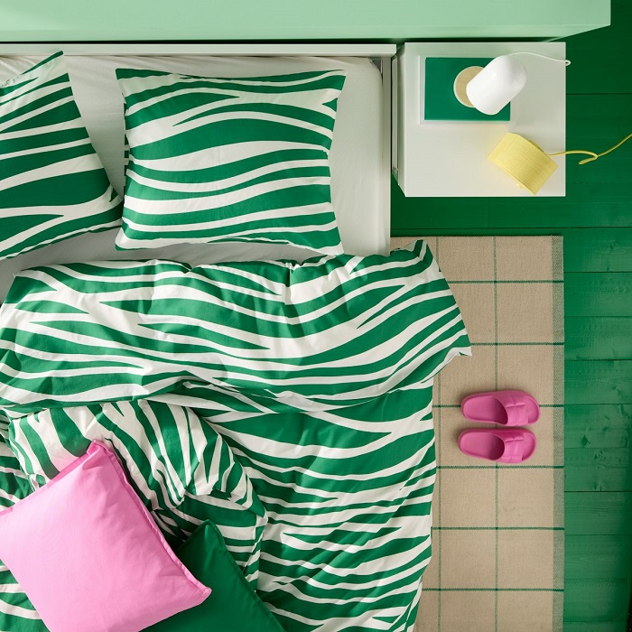 A PORTLAKMÅLLA duvet cover set with wavy green stripes, paired with a pink cushion and matching slippers, in a vibrant, green-themed room.
