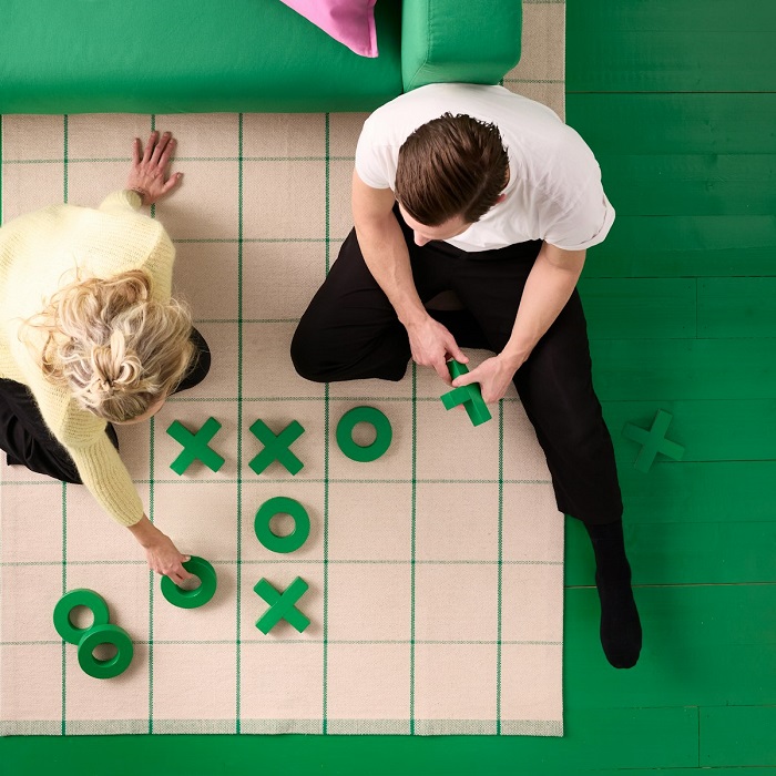 A RIDSTIG rug with a grid pattern, featuring two people playing a game of tic-tac-toe with green X and O pieces, on the floor next to a green sofa.