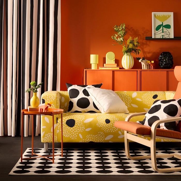 A yellow and white floral-patterned KLIPPAN cover for 2-seat sofa on a sofa in a vivid living room with an orange back wall.