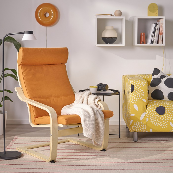 A bright orange POÄNG armchair with a cosy throw, placed next to a yellow sofa with a floral pattern, in a light and airy living room.