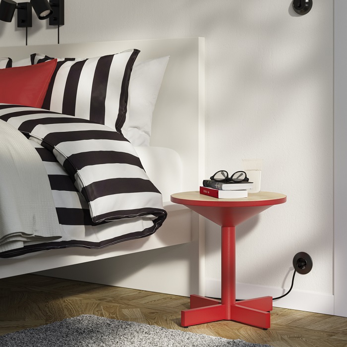 A red GRIMSARBO side table next to a bed featuring a red cushion and black-and-white striped bedding.