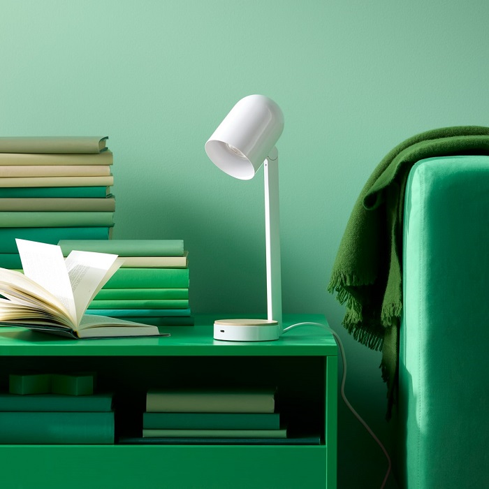 A white ENSMÄRKE table lamp on a green side table with books, beside a green sofa.