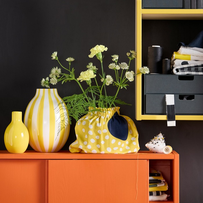 A white vase with yellow stripes sits on an orange cabinet with sliding doors, next to wild flowers placed in a yellow fabric bag.