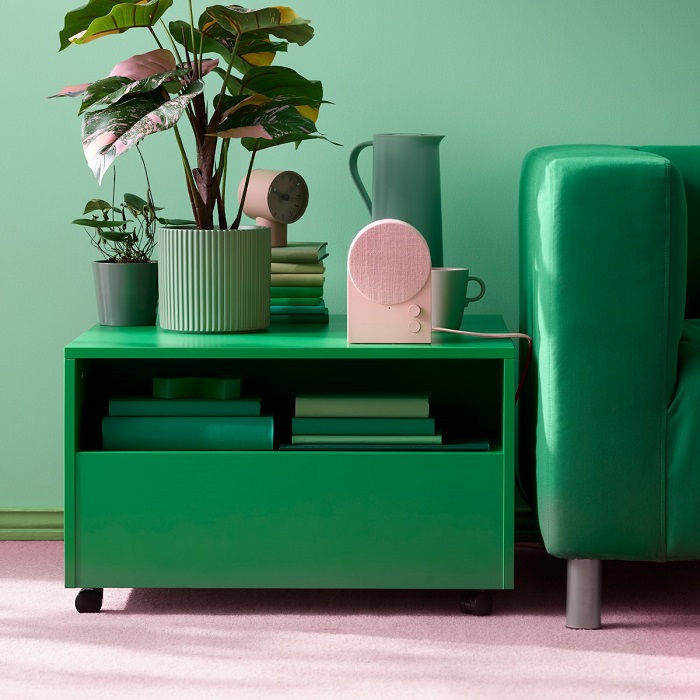 A green side table with a NATTBAD Bluetooth speaker, a potted plant and some green books, placed next to a green sofa in a pastel-coloured room.
