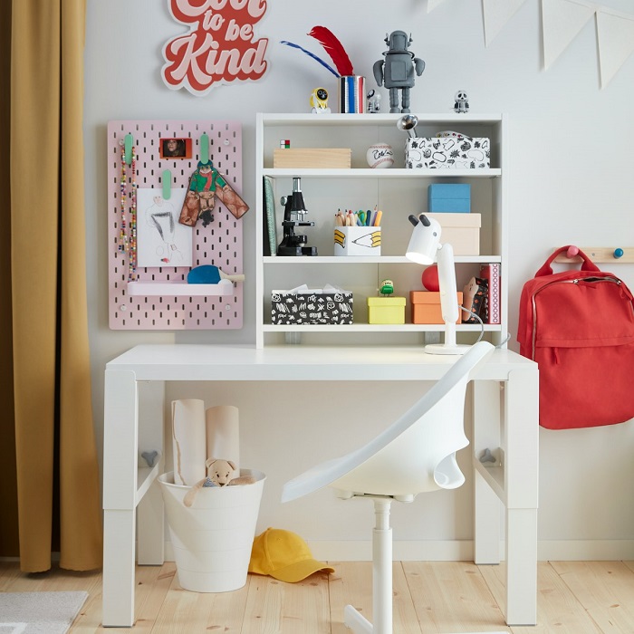 A child’s study corner with a white desk, chair, shelving unit filled with colourful stationery, and a red backpack hanging on the side.