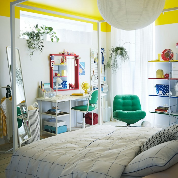 A bright children’s room with a white desk, green swivel chair, red-framed mirror, hanging plants, and a green armchair.