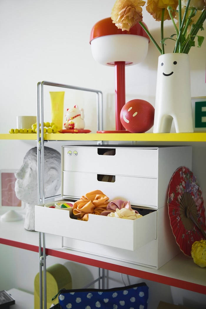 Two white mini chests stacked together with a total of four drawers—one slightly open—sitting on a colourful shelf alongside a red table lamp and a smiling vase.