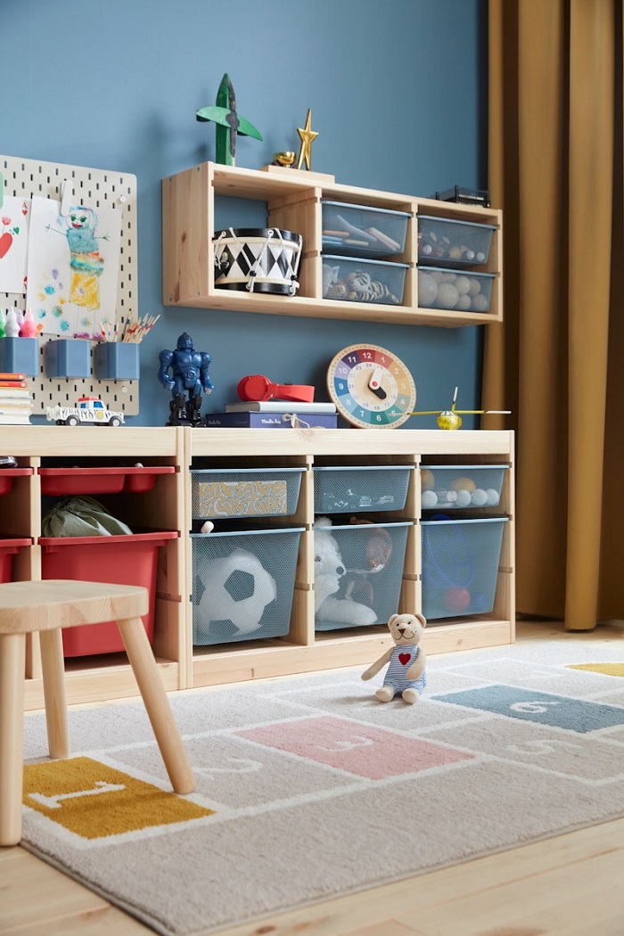 A wooden storage unit with mesh boxes placed against a blue wall, with a colorful hopscotch-patterned rug spread out on the floor in front.