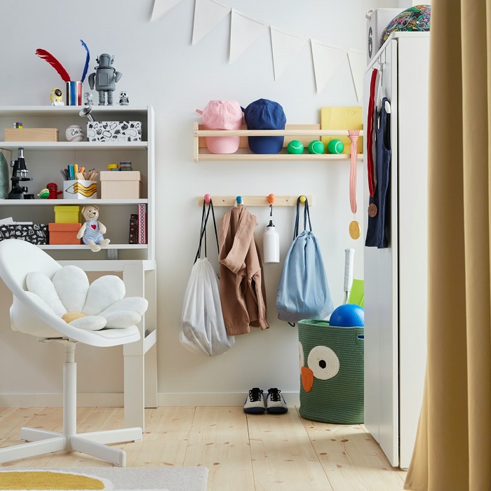 A corner of a children’s room featuring a white desk with a matching chair, open wall shelves holding books, and wall hooks displaying hanging clothes.