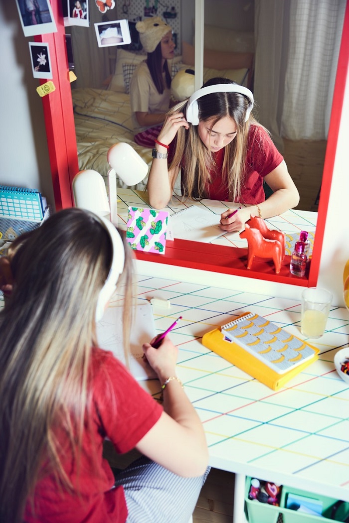 A teen wearing headphones is sitting at their desk in front of a mirror with a red frame, doing homework.