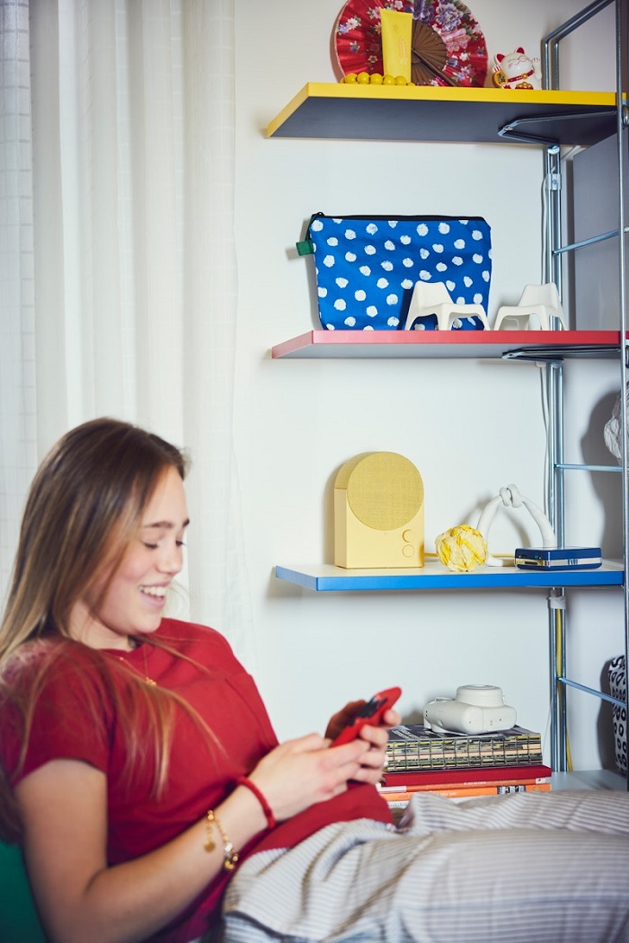 A teen sitting by the wall, looking at their smartphone, with a colourful open shelving unit in the background.