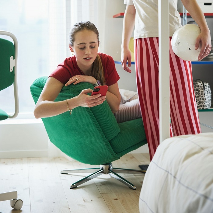 A teen sits on a low green armchair looking at their smartphone, while another teen stands nearby.