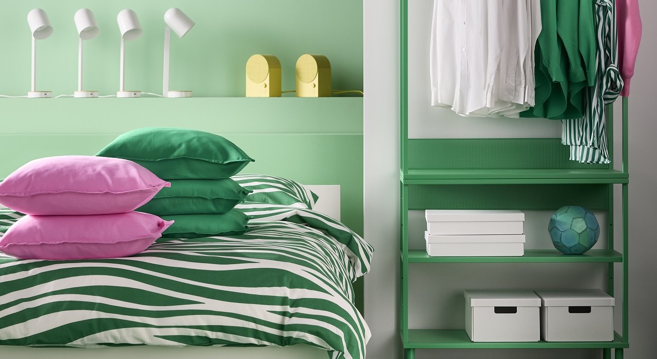 A vibrant bedroom with a green and white striped bedding set, featuring pink and green cushions, an open shelving unit in green, fourmodern white table lamps and two yellow speakers.