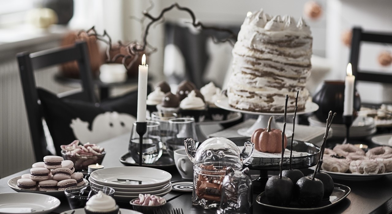 A dark table with Halloween treats served on KUSTFYR side plates, along with a KUSTFYR bowl with a lid shaped like a ghost and some lit candles.