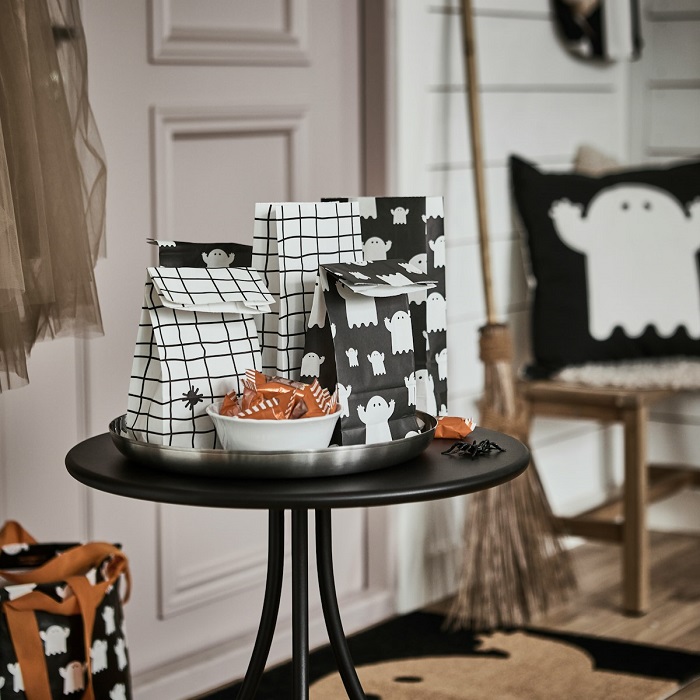 Some KUSTFYR paper bags featuring ghosts and spiders lie on a small black table in a hallway, next to a bowl full of treats.