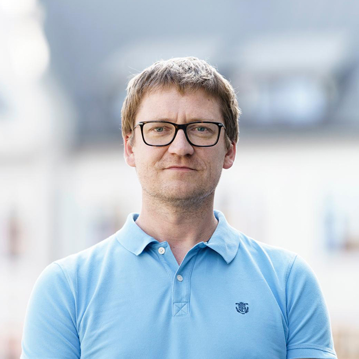 Vīrieša portrets gaiši zilā polo kreklā ar brillēm, ārpus telpām.
