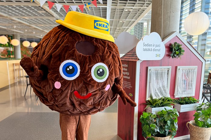 Cilvēks lielā, apaļā, brūnā gaļas bumbiņas kostīmā ar lielām acīm, sarkanu muti un rozā vaigiem, dzeltenā IKEA cepurē stāv IKEA veikalā blakus sarkanai rotaļu mājiņai ar augiem un zīmi.