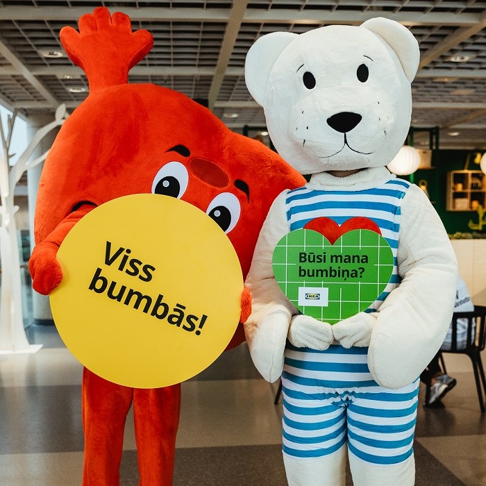 Divi cilvēki sarkanā sirds un baltā lāča kostīmā stāv kopā IKEA veikalā, turot zīmes ar uzrakstu