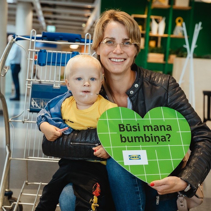 Smaidoša sieviete tur klēpī mazu bērnu IKEA veikalā. Viņi tur zaļu zīmi ar uzrakstu "Būsi mana bumbiņa?"